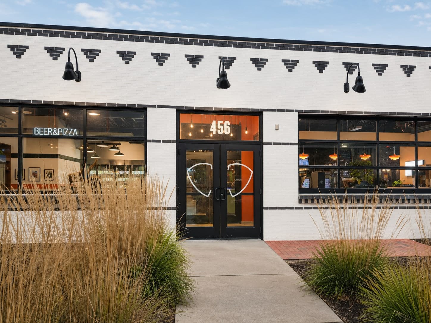 Entry doors at Elephant & Co. framed in white brick with a black corbelled frieze, gooseneck lamps, and ornamental grasses