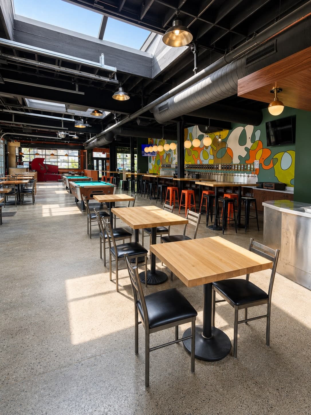 Skylit dining room with butcher-block tables, orange stools, the self-pour tap wall, and the full-height mural